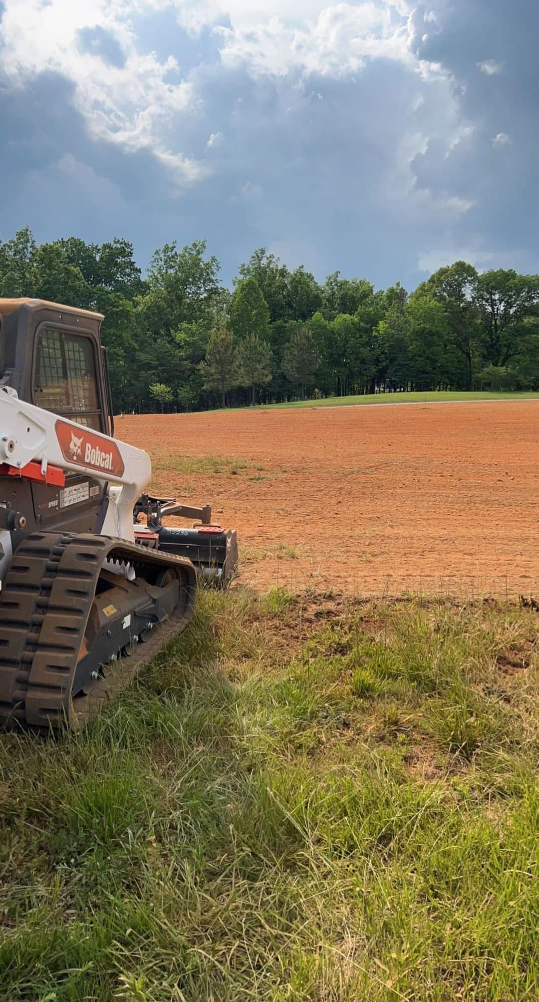 Bobcat skid steer equipment on freshly tilled soil, surrounded by lush green trees.