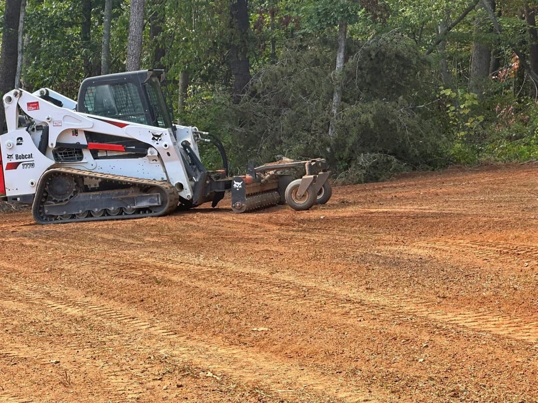 Bobcat skid steer with brush cutter clearing land in a forested area.