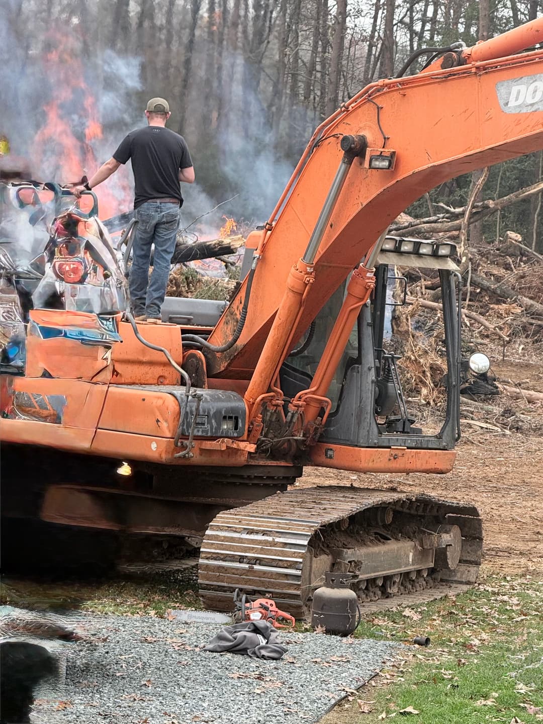 Person operating an orange excavator with smoke from nearby fire and forest debris in background.