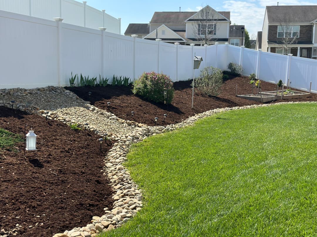 Landscaped backyard featuring rocks, flower beds, and decorative lighting against a white fence.