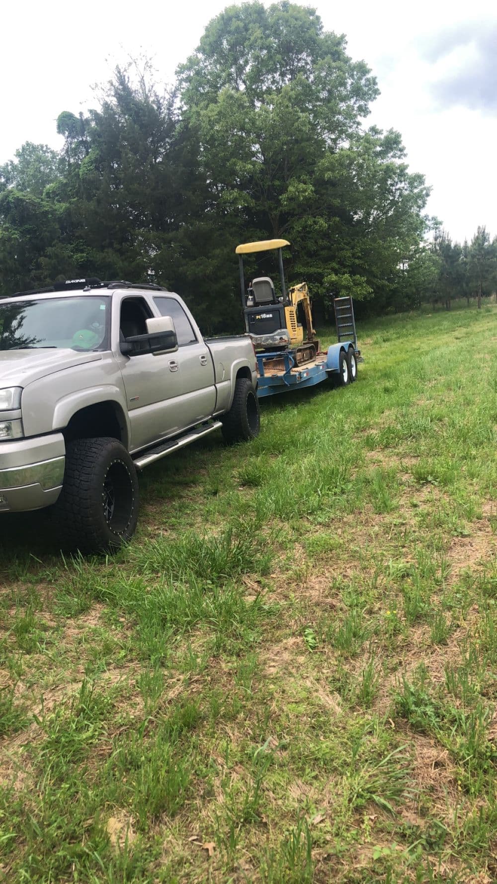 Silver pickup truck towing a flatbed trailer with construction equipment in a grassy field.