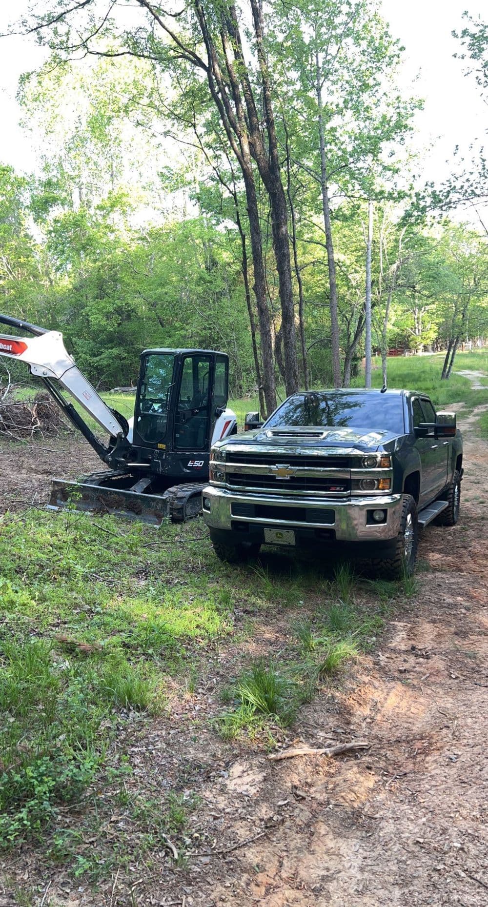 Chevrolet truck parked beside a compact excavator in a wooded area.