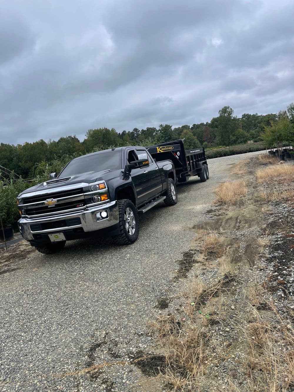 Black Chevrolet Silverado truck pulling a dump trailer on a gravel road surrounded by trees.