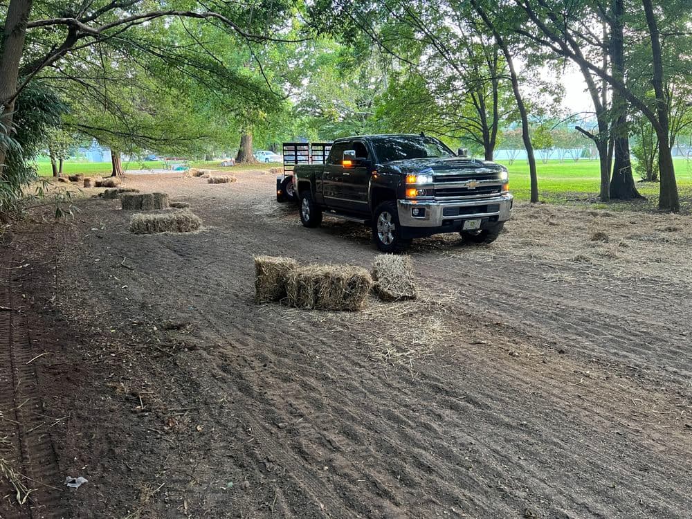 Black pickup truck with hay bales parked on a dirt road lined with trees in a rural setting.