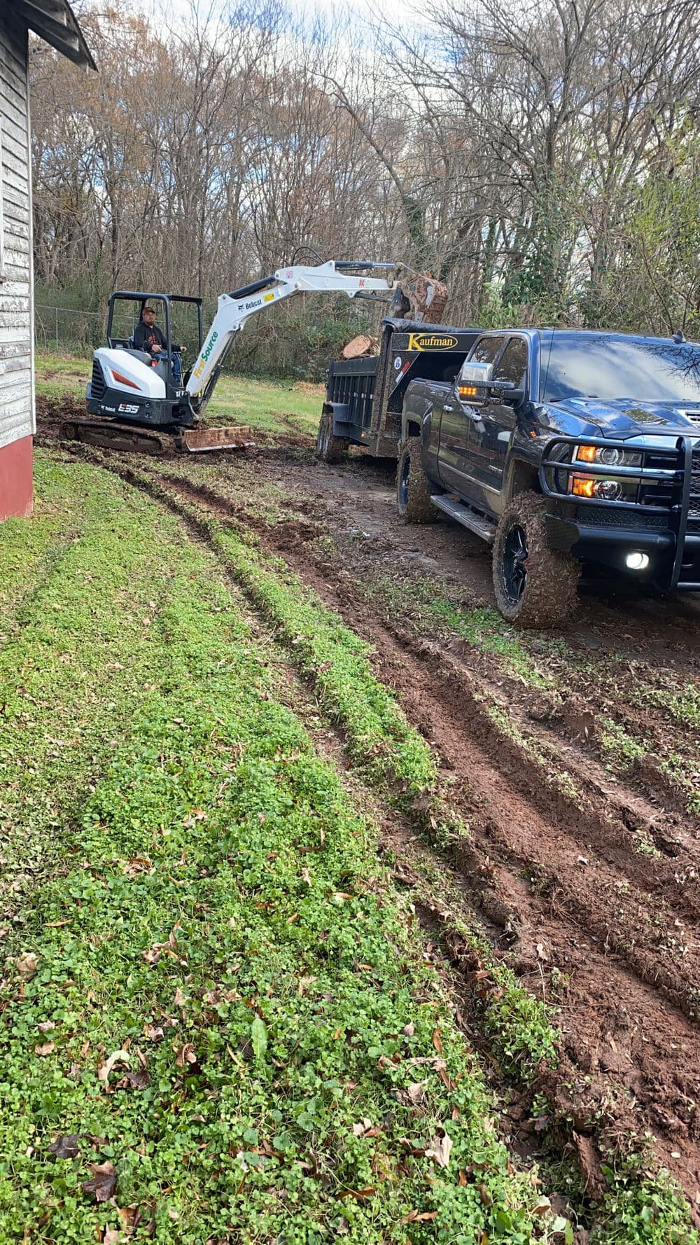 Excavator digging near a house with a truck and trailer in a muddy yard.