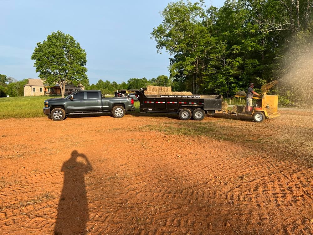 Pickup truck with trailer unloading hay bales on an orange dirt field surrounded by trees.