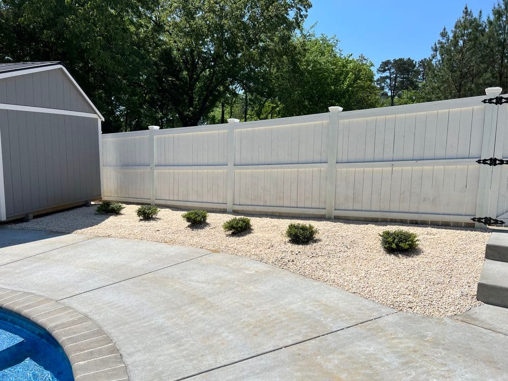 White vinyl fence with landscaped gravel area and small shrubs near a swimming pool.