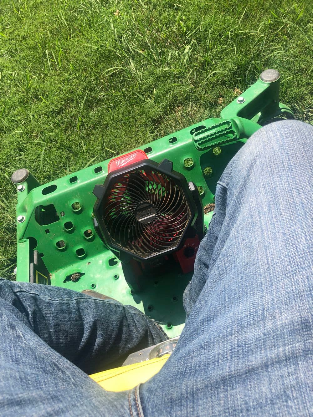 Person sitting on a green lawnmower with a Milwaukee fan on their lap, surrounded by grass.