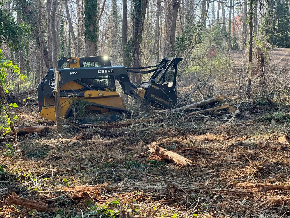 John Deere 331G land clearing machine working in a wooded area, clearing fallen trees and brush.
