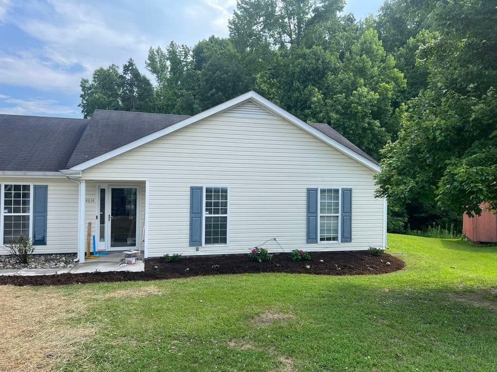 Charming white house exterior with blue shutters and landscaped yard, surrounded by trees.