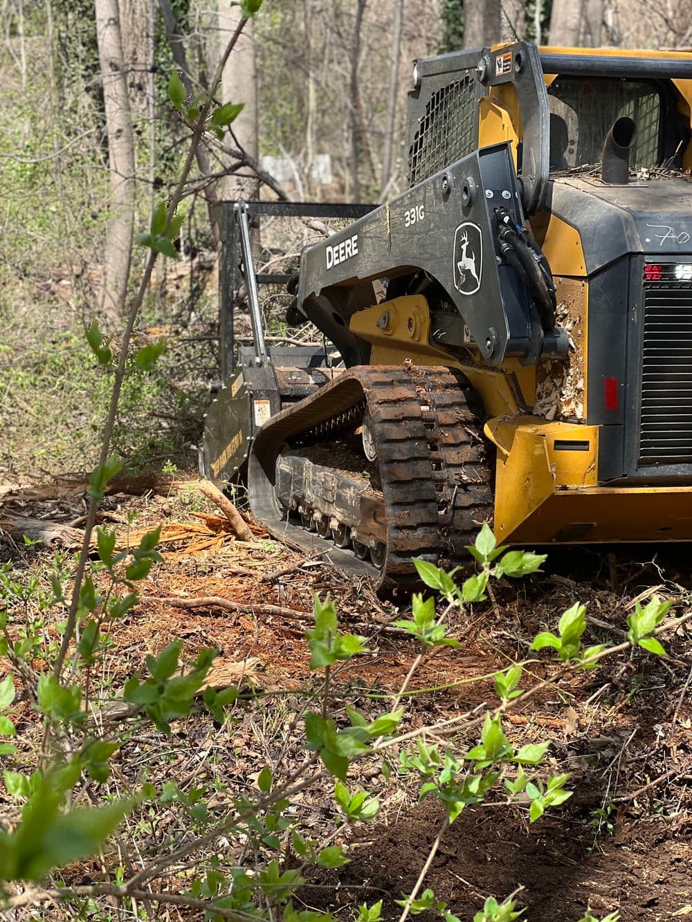 John Deere 331G cab track loader clearing vegetation in wooded area with fresh green leaves.