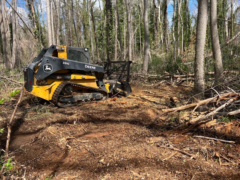 John Deere 331G forest machine clearing brush in wooded area, showcasing forestry work.