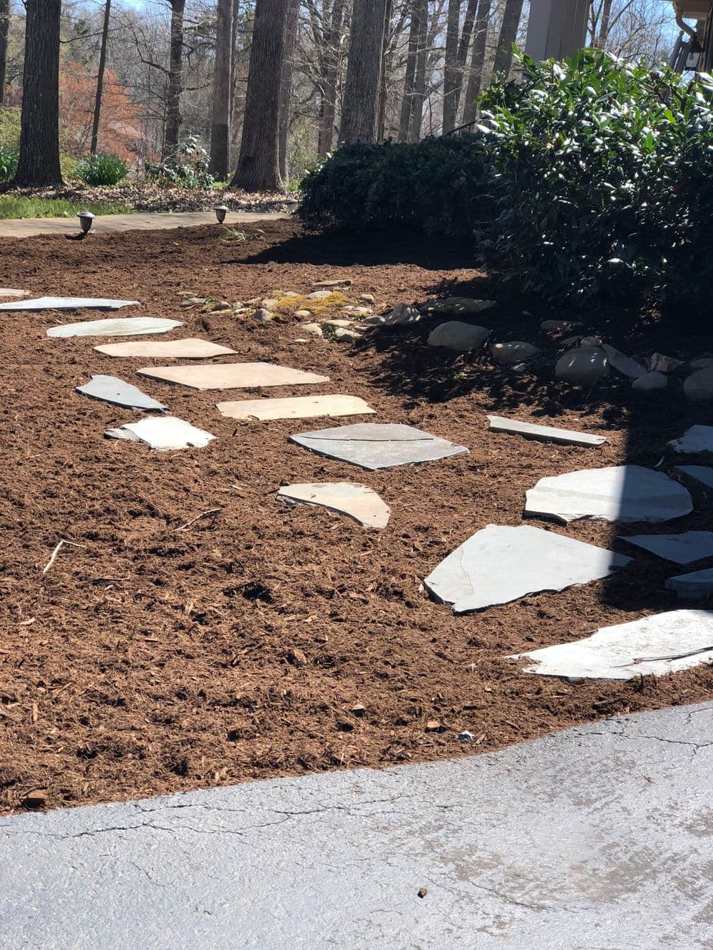 Pathway made of stone slabs through freshly mulched garden and trees in the background.