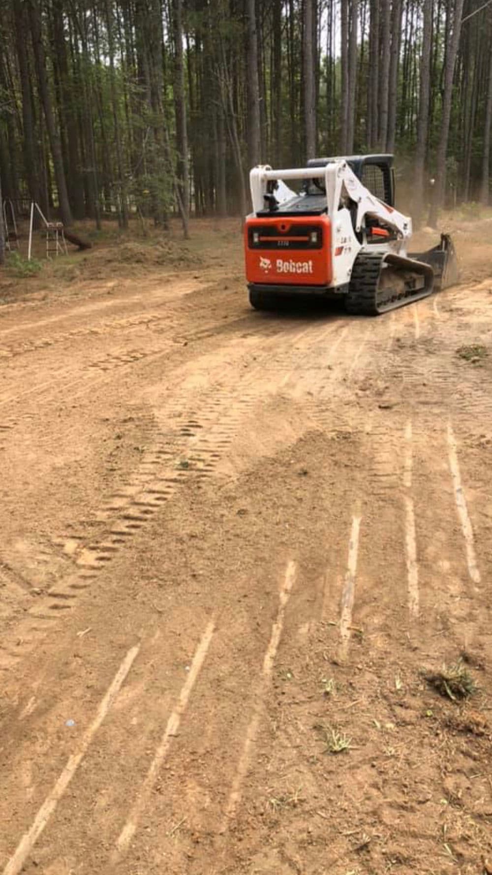 Bobcat skid steer clearing dirt in a wooded area with visible tire tracks on the ground.